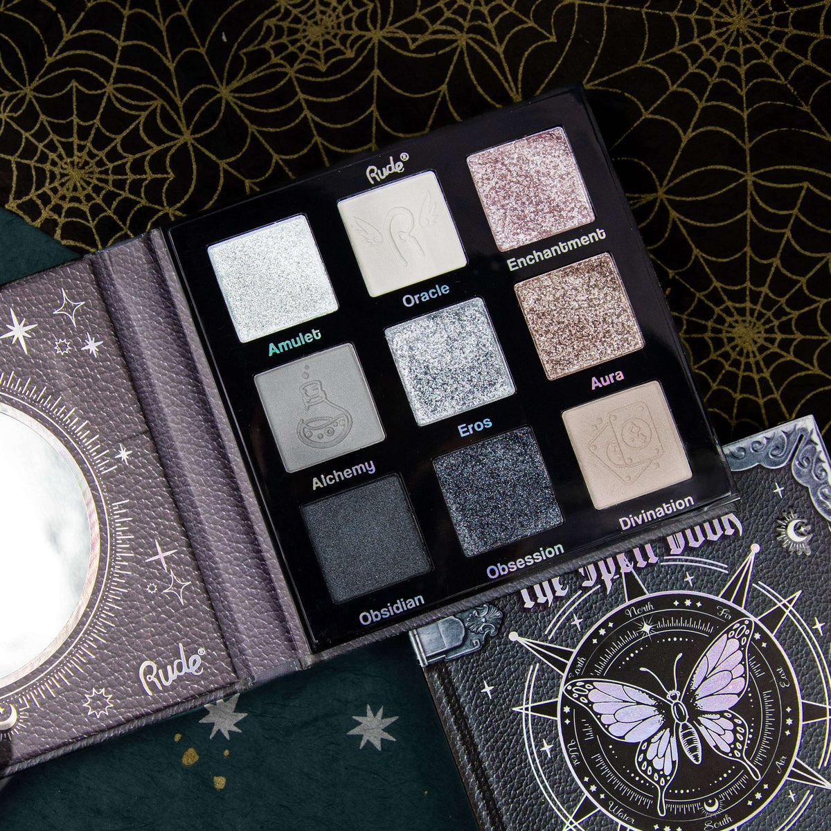 An open Rude brand eyeshadow palette displaying nine shimmering shades of black and grey labeled Amulet, Oracle, Enchantment, Alchemy, Eros, Aura, Obsidian, Obsession, and Divination, styled on a dark patterned background.