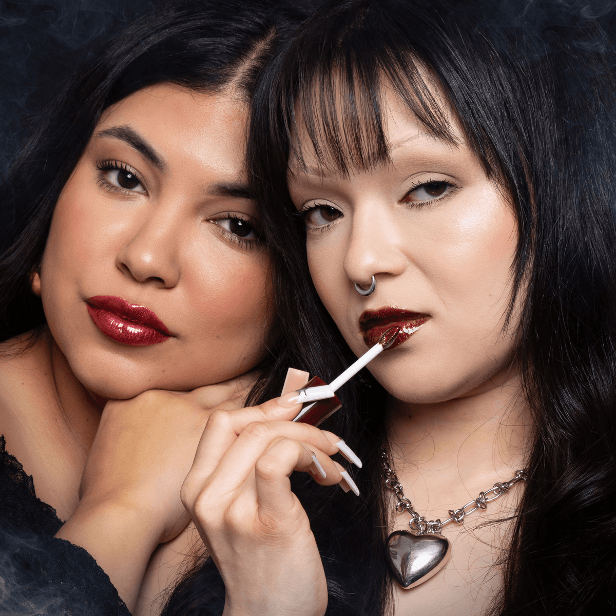 Two women applying red lip oil with a dark background.