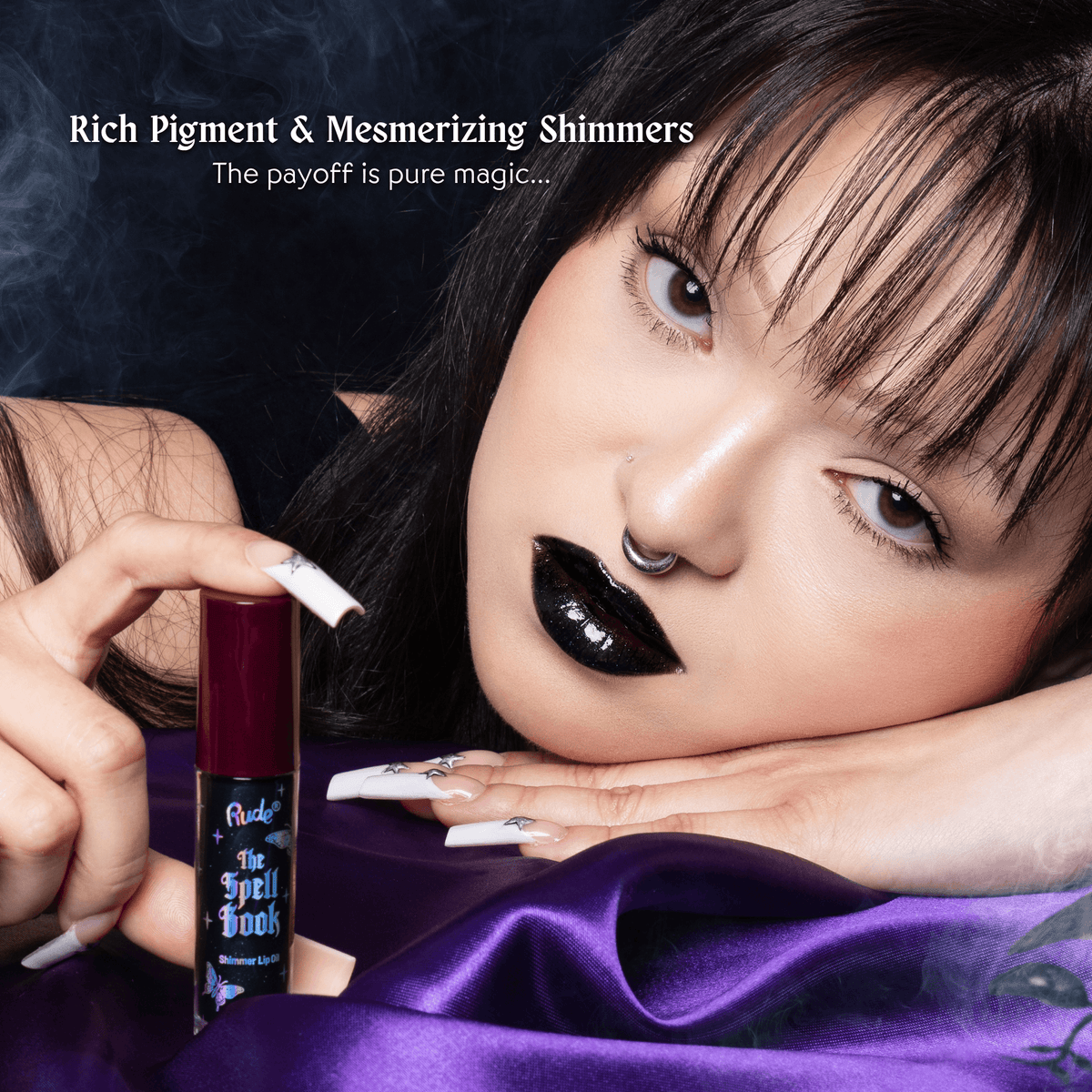 A close-up lifestyle shot of a model with black lipstick and white nails holding a tube of lip oil. The model is resting her head on her hand, draped in purple satin, with dark smoke in the background and text that reads &