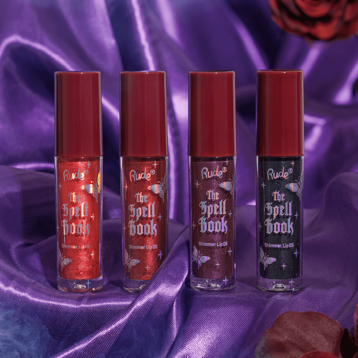 Four lip gloss bottles with red caps and iridescent labels on a purple fabric background. The lip glosses are various shades of red to black and shimmer in the lighting.