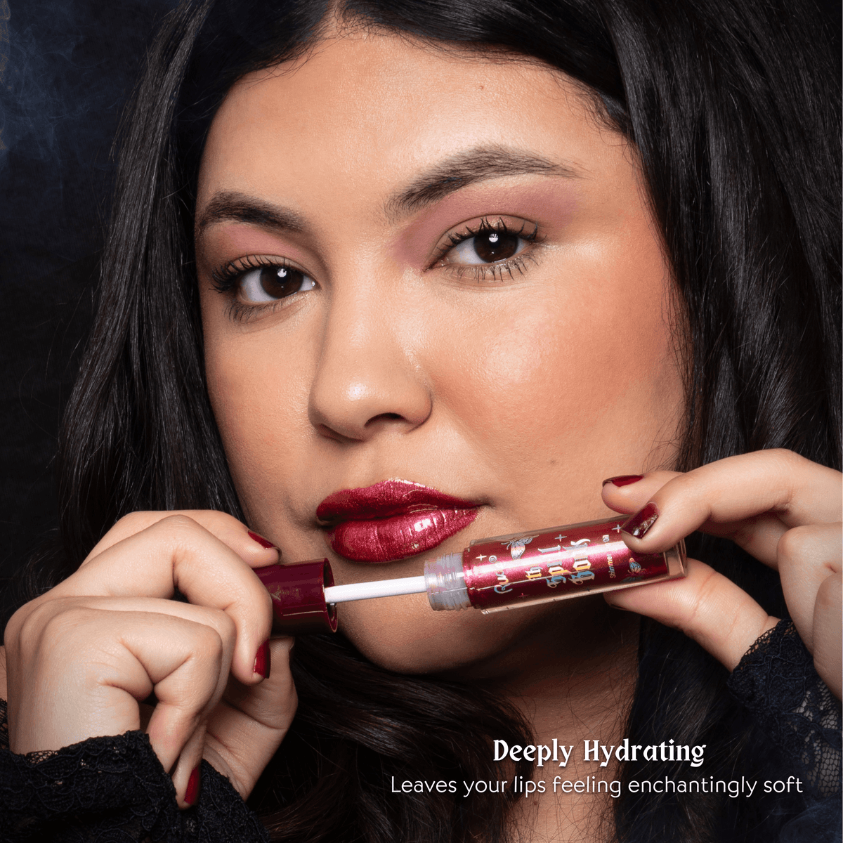 A close-up of a woman holding a tube of lip oil near her lips, showing the applicator. The lip oil tube is metallic red with a darker cap, and the text at the bottom reads &