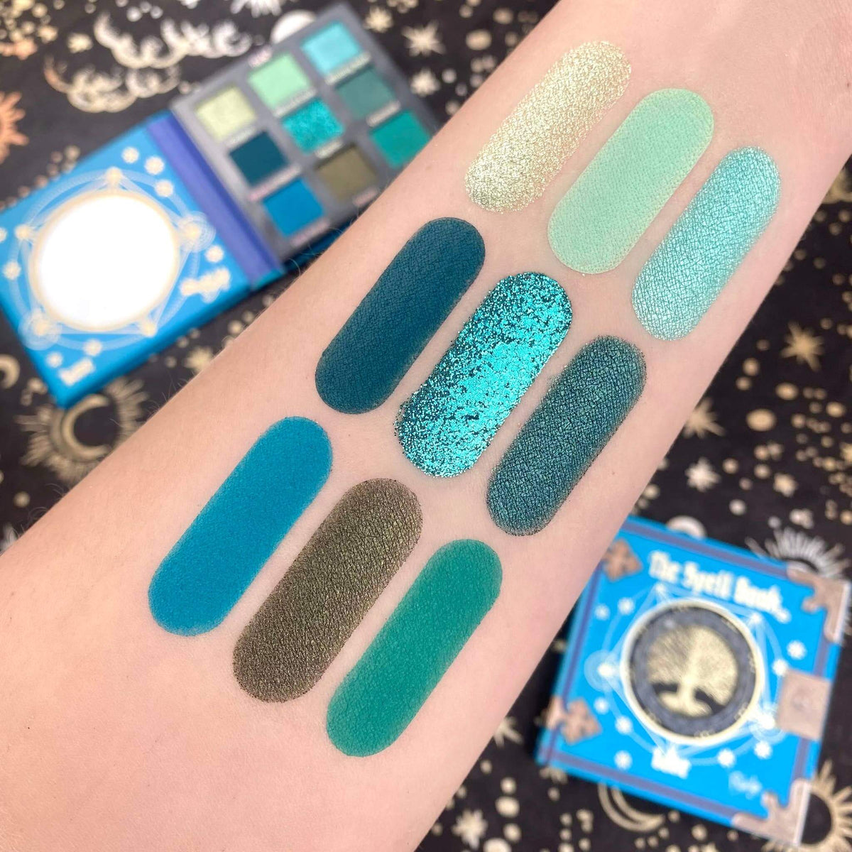 Blue color swatches on an arm with an open eyeshadow palette in the background.