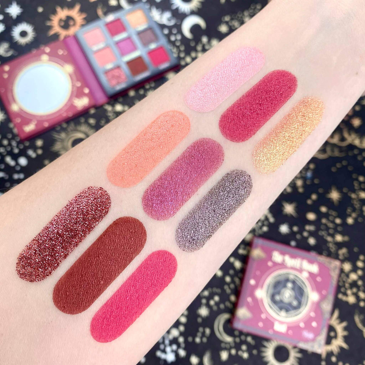 Pink and red makeup swatches on an arm with a colorful eyeshadow palette in the background.