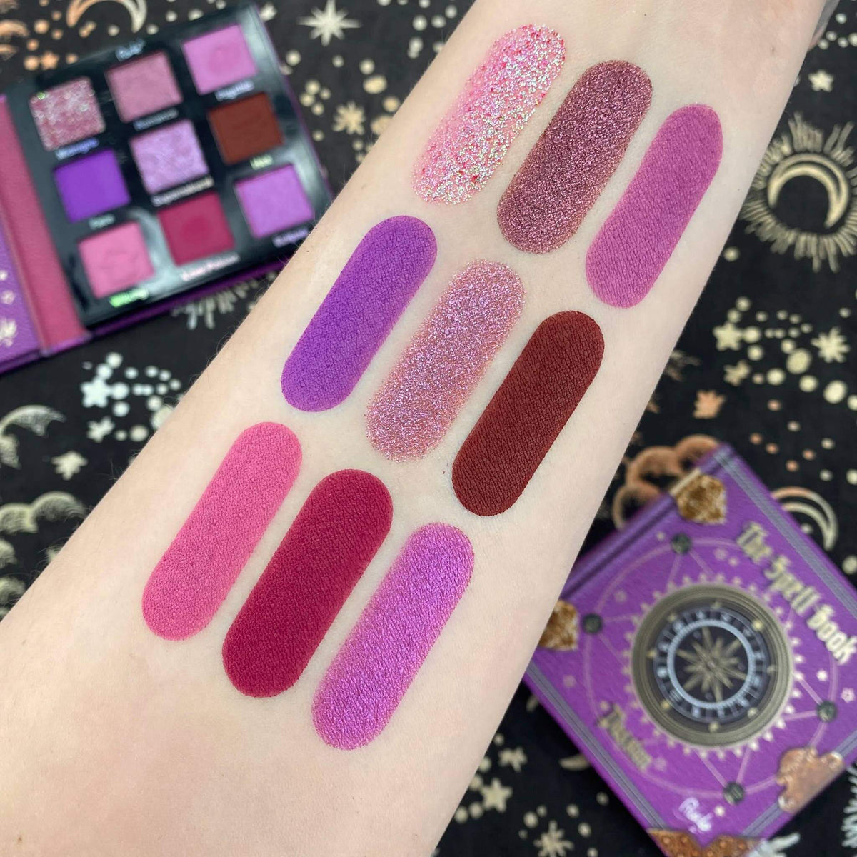 Pink, purple, and red eyeshadow swatches on an arm with an open eyeshadow palette in the background.