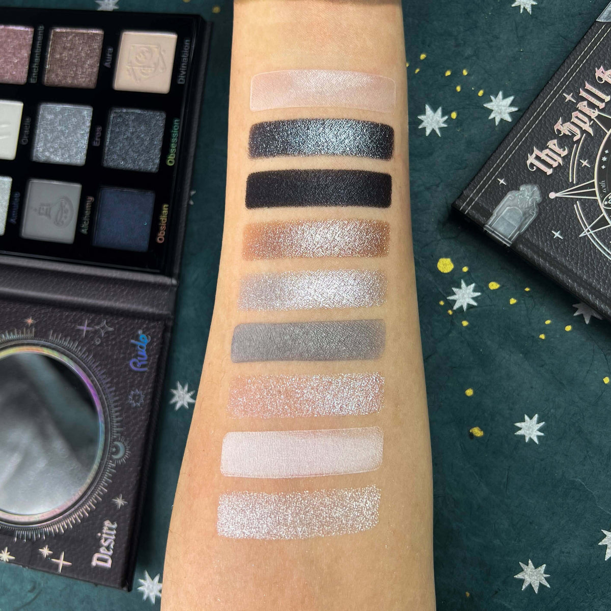 An arm with nine vertical swatches of shimmer eyeshadow in neutral and metallic shades, shown next to an open eyeshadow palette.