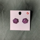 Purple polyhedral dice earrings on a pink card against a green background