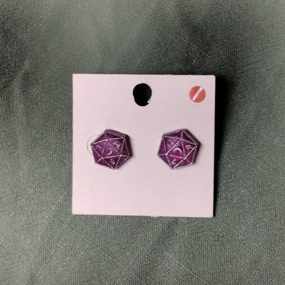 Purple polyhedral dice earrings on a pink card against a green background